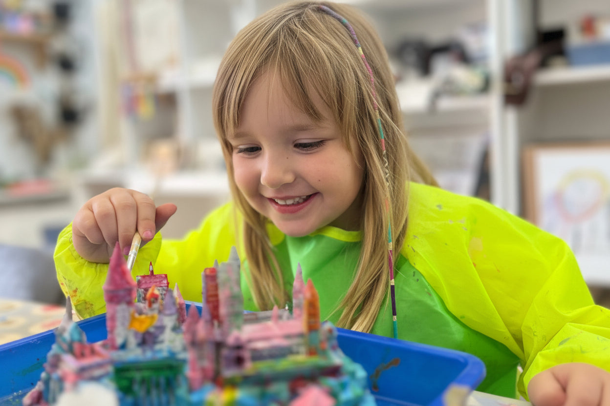 Child painting a toy castle in a colorful indoor setting, the wild fig and fern stay & create