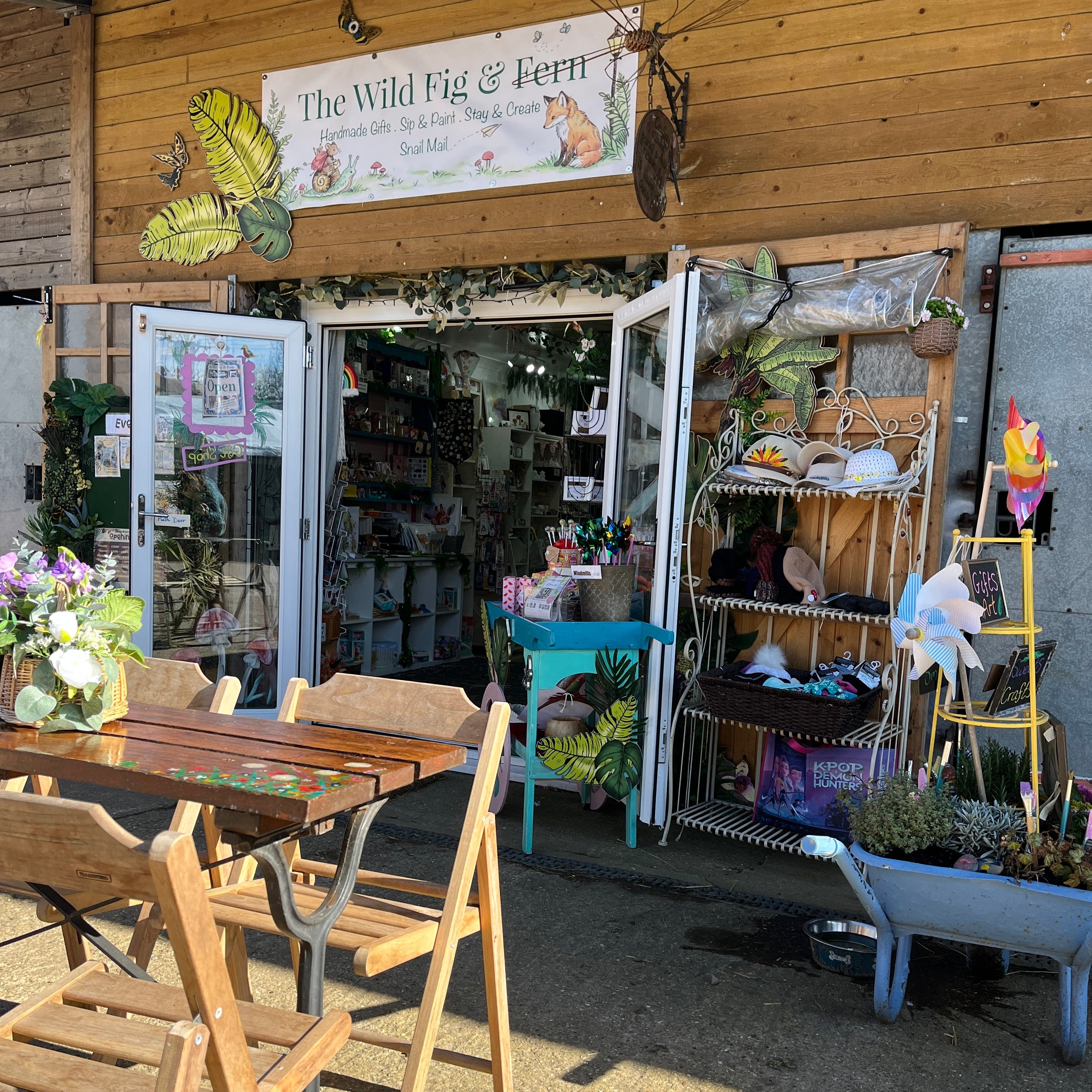 Storefront with outdoor seating area featuring a wooden table and chairs, surrounded by plants and decorative items.